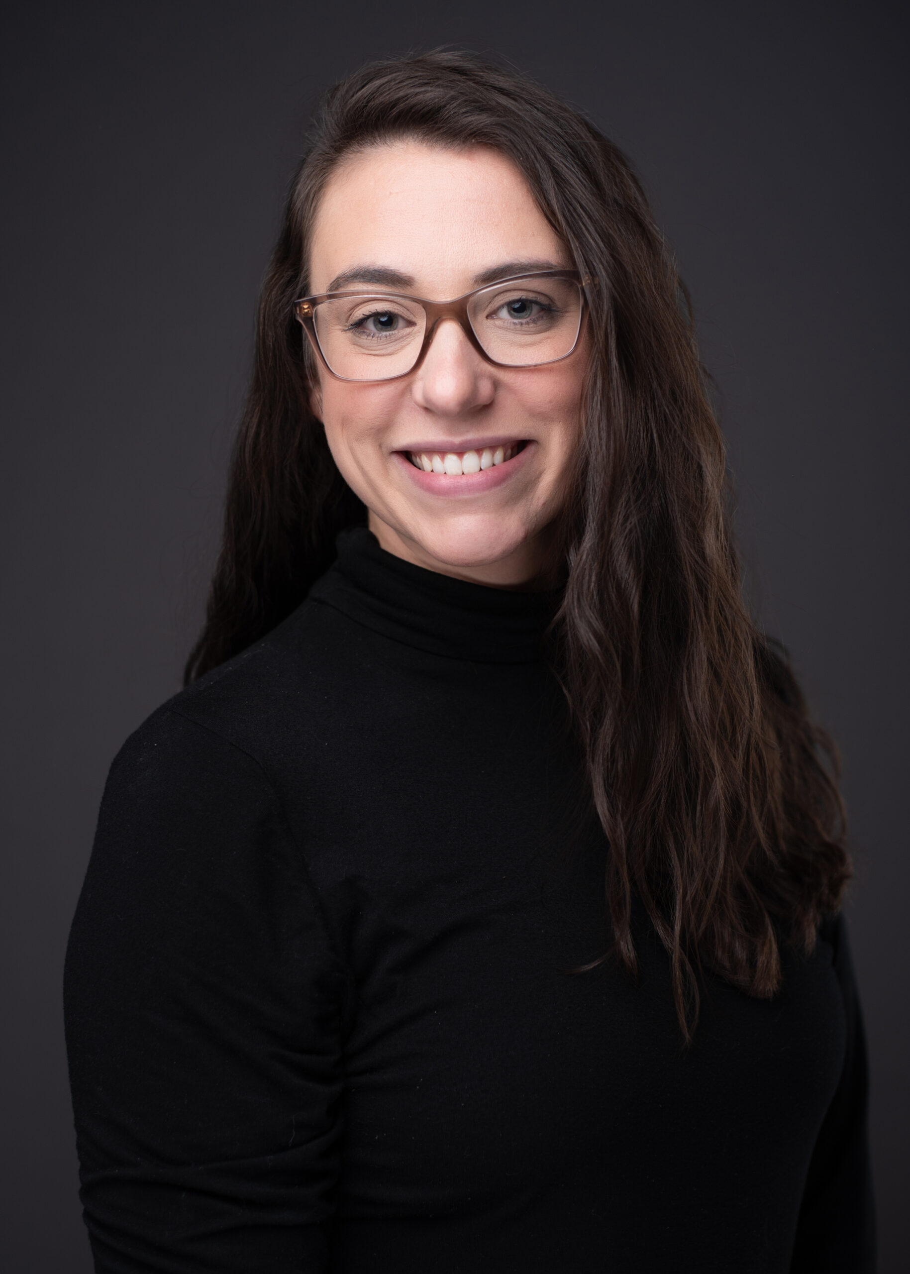 Portrait of a smiling woman with long dark hair and glasses wearing a black turtleneck against a dark background.