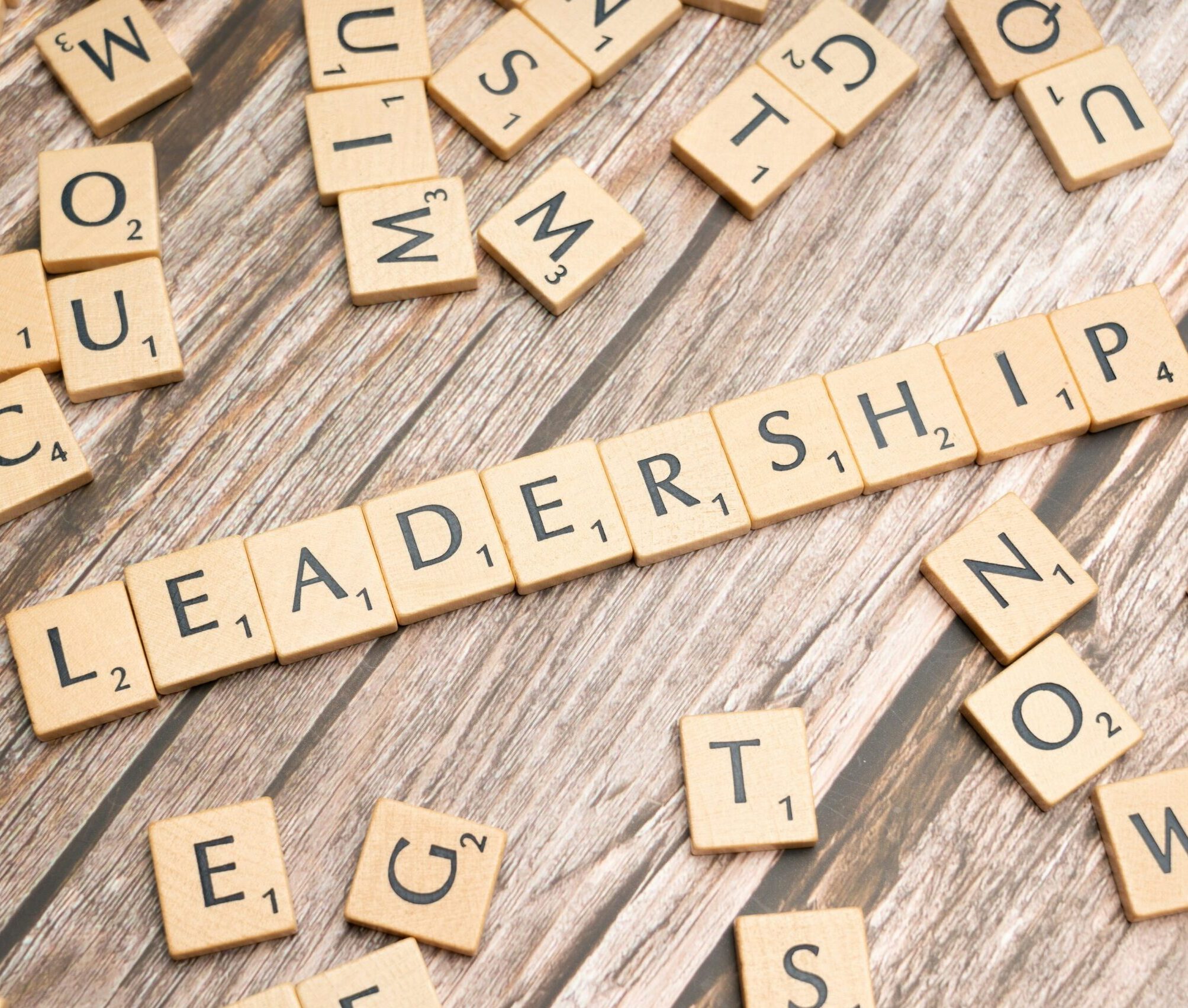 Close-up of Scrabble tiles on a wood surface forming the word 'LEADER' with other scattered letters nearby.
